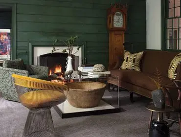 Living room with dark green paneled walls, fireplace, grandfather clock, brown sofa with patterned pillows, and a yellow wire chair.