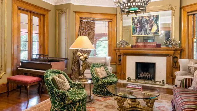 Living room with dark wood piano, ornate fireplace with a painting above the mantel, and green velvet armchairs with gold accents.