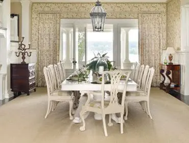Formal dining room with a long white table set for a meal, surrounded by upholstered chairs. Features include patterned wallpaper, floor-to-ceiling curtains, a dark wood buffet, and a built-in window seat.