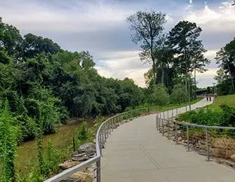 Curving paved pathway with metal railings alongside a wooded creek and green hillside under a cloudy sky
