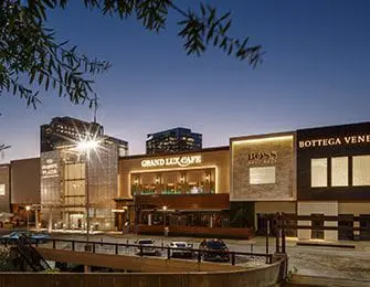 Modern shopping center at dusk with illuminated storefronts for Grand Lux Cafe, Boss, and Bottega Veneta.