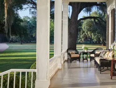 Southern estate porch with wicker furniture and flower arrangements, overlooking a manicured lawn and winding path under moss-draped oak trees.
