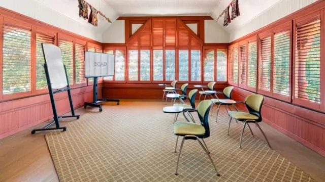 Meeting room with coral walls, wood shutters, a large screen displaying 'SCAD', a whiteboard, and modern chairs with yellow seats on a patterned carpet.