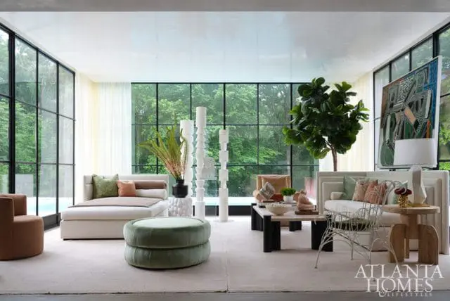 Living room with white sectional sofa, green tufted ottoman, and large windows overlooking greenery. Artwork and a fiddle leaf fig tree add decorative elements.