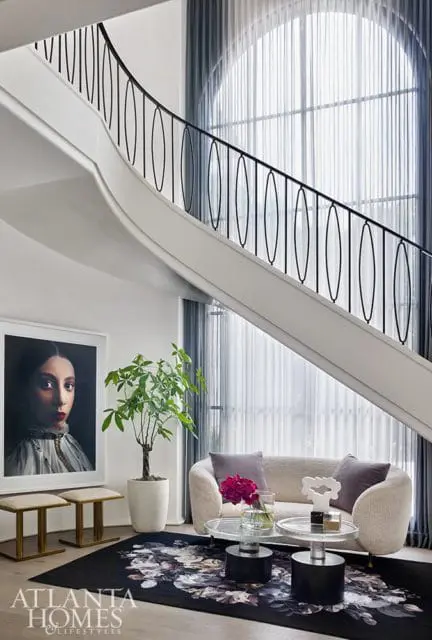 Living room with a curved white staircase, black railing, large arched window with sheer curtains, a boucle sofa, two glass coffee tables, and a floral rug.