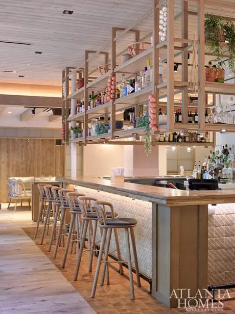 Bar area with light wood counter, wooden stools with dark seats, and shelves stocked with bottles and hanging garlic.