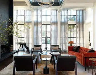 Living room with a tufted orange sofa, dark wood armchairs, and floor-to-ceiling windows with patterned sheer curtains. A mirrored ceiling reflects a modern chandelier.