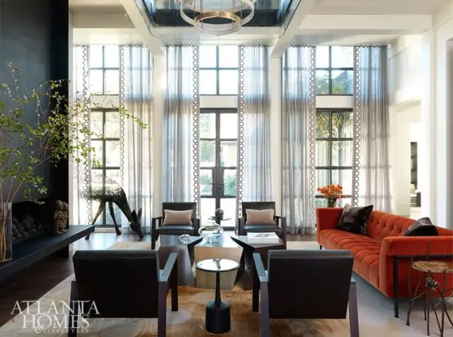 Living room with floor-to-ceiling windows draped in sheer curtains, a black fireplace, dark leather armchairs, and a vibrant orange tufted sofa.