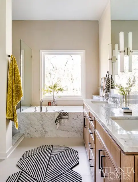 Modern bathroom with a marble-clad soaking tub, wood vanity with marble countertop, and a graphic black and white rug.