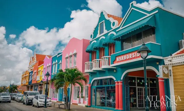 Row of colorful buildings in turquoise, pink, and yellow with arched doorways and balconies, parked cars on the street, and palm trees under a blue sky with clouds.