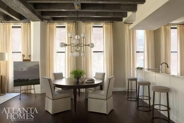 Loft dining area with dark wood floors, exposed beams, cream curtains, a round dining table with upholstered chairs, and a modern globe chandelier. A kitchen island with bar stools is visible to the right.