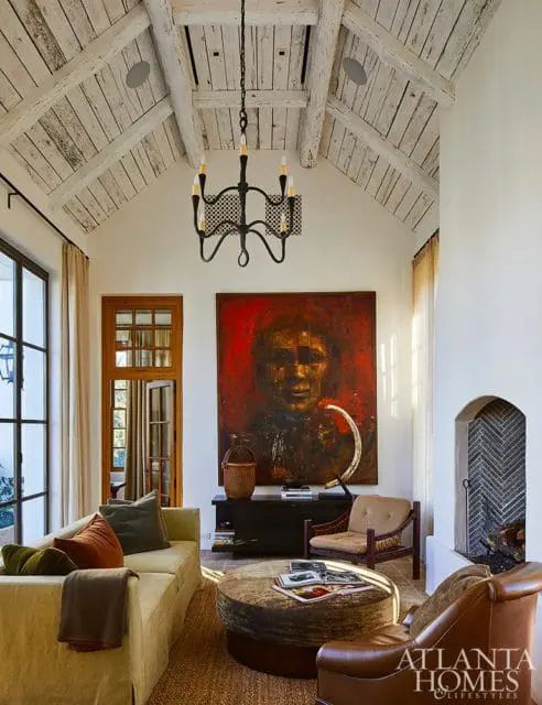 Living room with vaulted, whitewashed wood-beamed ceiling, large abstract portrait, neutral sofa, round ottoman coffee table, and fireplace with herringbone tile surround.