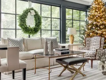 Living room with a large Christmas tree, a green wreath on the window, a neutral sofa, patterned accent chairs, and a coffee table with books.