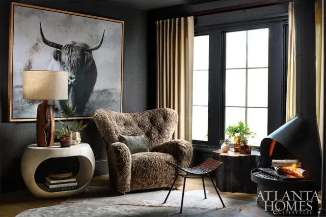 Dark gray living room with a large black and white Highland cow print above a textured brown armchair, a sculptural side table, and a modern black fireplace with burning logs.