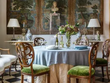 Dining room with a round table covered in a light blue tablecloth, set with white plates, wine glasses, and bottles. Green velvet upholstered chairs surround the table, and a large tapestry hangs on the wall behind.