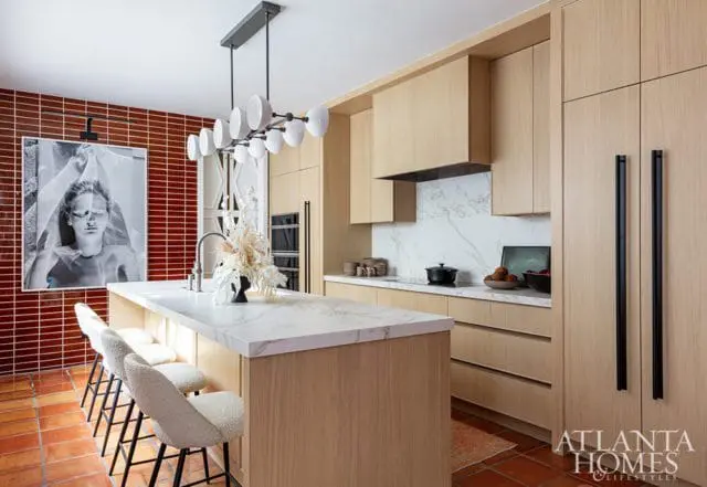 Modern kitchen with light wood cabinetry, white marble island and countertops, red tile backsplash, and a large black and white artwork.
