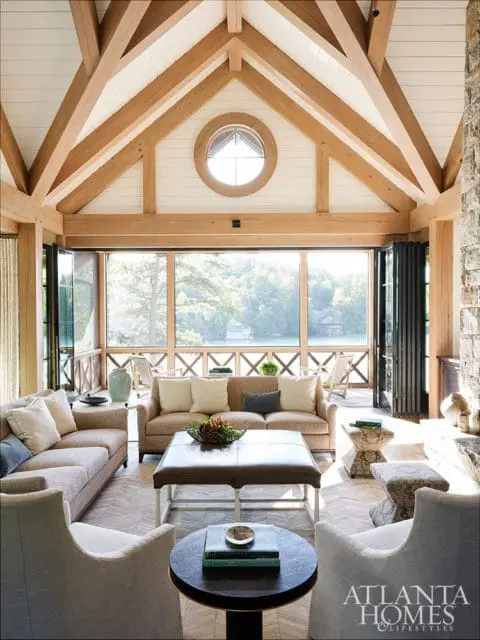 Living room with vaulted wood beam ceiling, stone fireplace, neutral sofas, leather ottoman coffee table, and screened porch overlooking a lake
