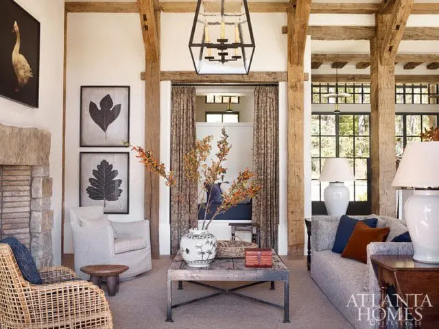 Living room with exposed wooden beams, stone fireplace, upholstered chairs, a sofa with throw pillows, and a coffee table with a vase of branches.