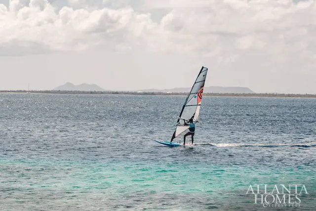 Windsurfer on a blue board with a white and black sail on the ocean, with distant islands under a cloudy sky. Atlanta Homes & Lifestyles logo in the lower right.