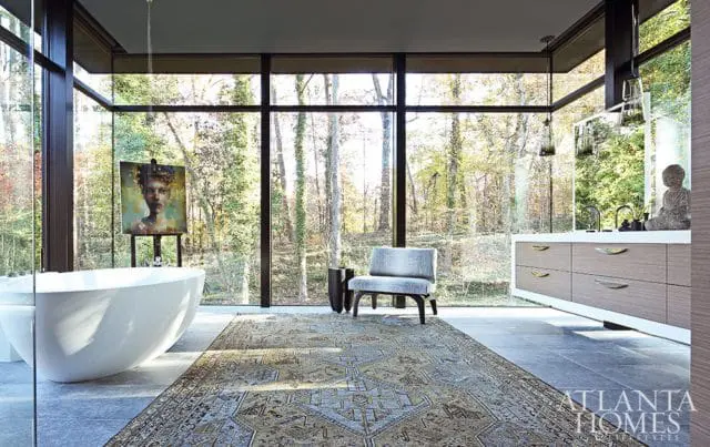 Modern bathroom with freestanding tub, artwork, and wood vanity overlooking a forest through floor-to-ceiling windows.