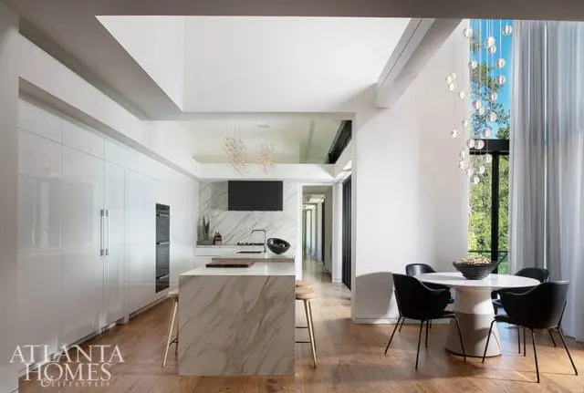 Modern white kitchen with marble island, integrated appliances, and dining area with black chairs.