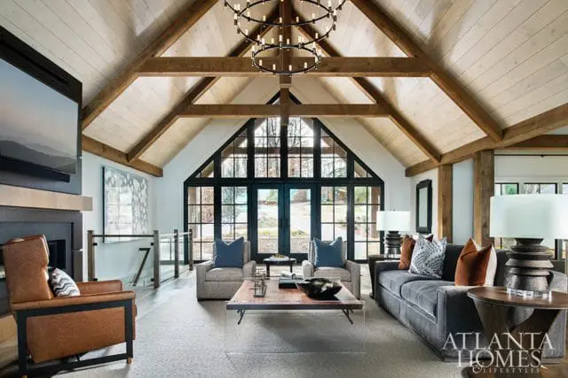 Living room with vaulted wood-beamed ceiling, large black-framed windows, gray sofa, leather armchair, and a rustic coffee table.
