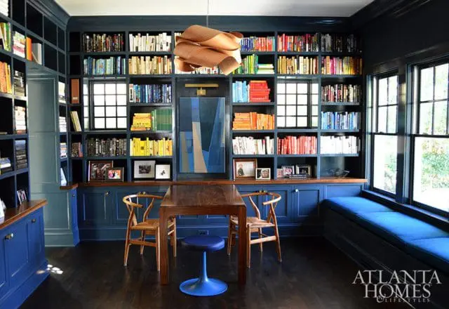 Dark blue library with floor-to-ceiling bookshelves, a wooden table with chairs, and a blue window seat.