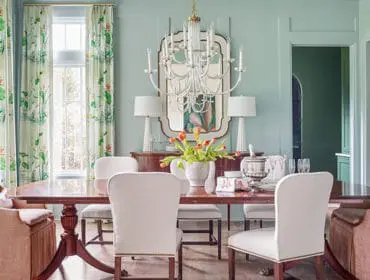 Light blue dining room with a dark wood table, upholstered chairs in cream and rust linen, botanical print curtains, and a crystal chandelier.