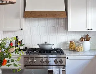 White kitchen with stainless steel gas range, elongated picket tile backsplash, and wooden range hood accent.