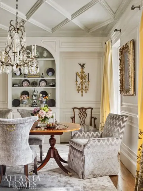 Dining room with a crystal chandelier, coffered ceiling, built-in shelves displaying decorative plates, and a round wooden table with upholstered chairs.