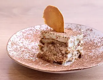 Tiramisu dessert dusted with cocoa powder on a white plate with a decorative cookie on top