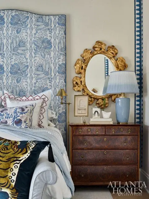 Bedroom with blue and white floral upholstered headboard, antique wood dresser, ornate gold mirror, and blue table lamp.