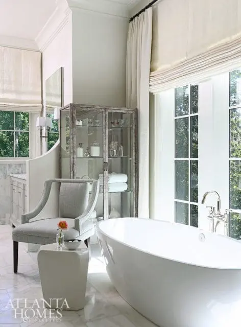 Master bathroom with a freestanding white bathtub, marble floors, a grey upholstered chair, and a glass-fronted cabinet filled with toiletries.
