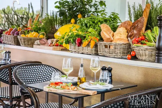 Table setting with wine glasses, olive oil dispenser, and pizza on a wooden board, with a display of fresh produce, bread, and charcuterie in wicker baskets in the background.