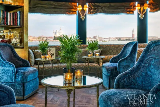 Lounge area with plush blue velvet armchairs and patterned sofa, overlooking a city skyline with domed buildings and a bell tower across a body of water.