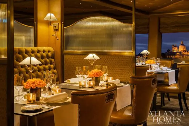Restaurant dining room with tufted gold banquettes, leather chairs, and tables set with white linens, orange roses, and candles. A cityscape view is visible through the windows.