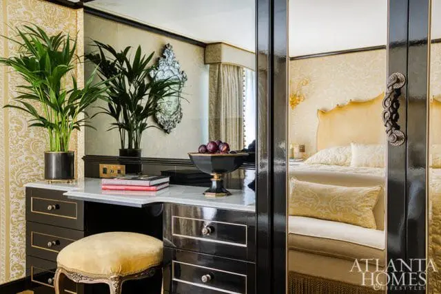 Black vanity with marble top, gold damask wallpaper, and a yellow upholstered stool. A mirrored closet reflects a bed with a gold headboard.