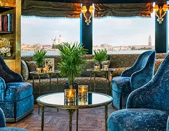 Lush blue velvet armchairs and sofa with ornate patterned upholstery in a room with large windows overlooking Venice, Italy.
