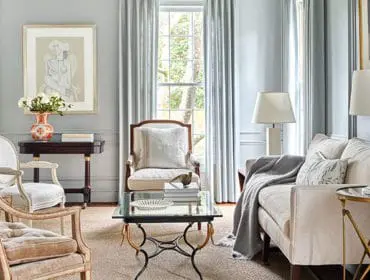 Light blue living room with upholstered chairs, a sofa, a glass-top coffee table, and a dark wood console table with a vase of flowers.