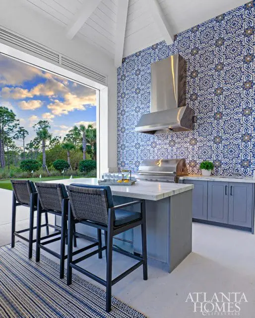 Outdoor kitchen with blue and white patterned tile backsplash, marble island with bar stools, and a large open window overlooking a golf course.