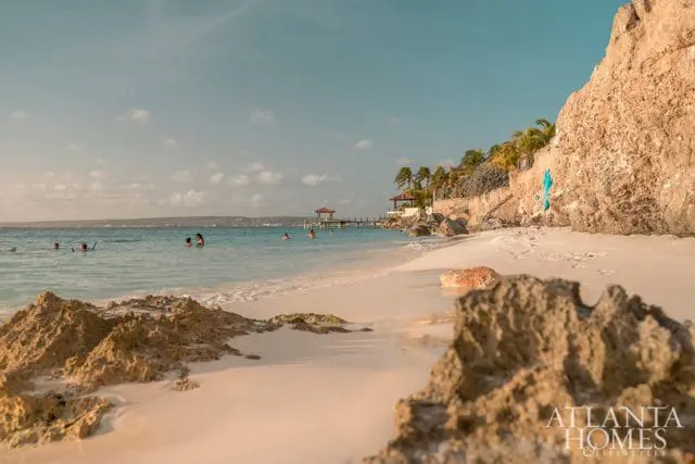 Sandy beach with turquoise water and people swimming. A stone cliff face with palm trees and a pier with gazebos is visible in the background.