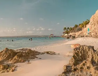 Sandy beach with rocky outcrops and turquoise water, people swimming in the distance, palm trees and a pier under a clear blue sky.
