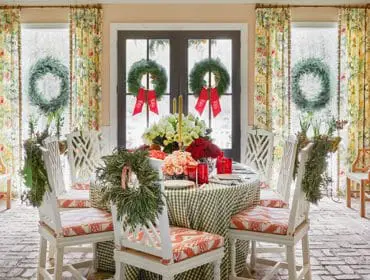 Dining room with a round table set for a holiday meal, featuring a green gingham tablecloth, floral curtains, and wreaths on the doors.