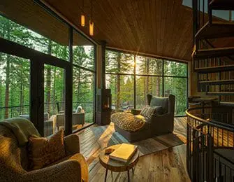 Living room with floor-to-ceiling windows overlooking a forest, featuring a spiral staircase, wood-paneled ceiling, and comfortable seating.
