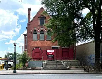Red brick historic building with arched windows and red shutters, featuring an "Available" sign on the front steps.