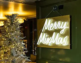 A gold sign with white neon text reading "Merry Mixmas" hangs on a dark green door next to a silver Christmas tree decorated with warm white lights.