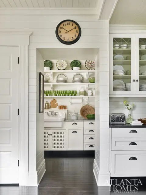White kitchen with shiplap walls, a butler's pantry with open shelving displaying plates and glasses, a farmhouse sink, and white cabinetry. A round clock hangs above the pantry.