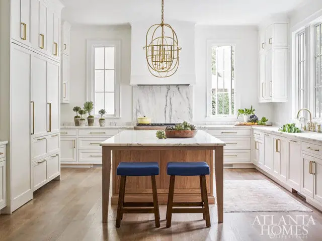 White kitchen with marble countertops, custom white cabinetry with brass hardware, and a wooden island with blue stools.