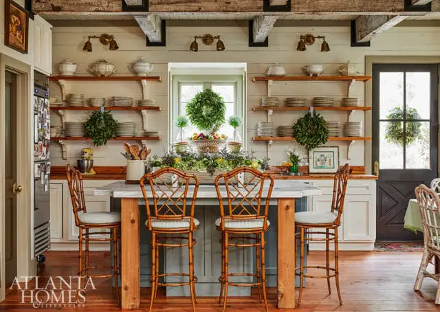 Farmhouse kitchen with open shelving displaying white dishes, a marble-topped island with bamboo stools, and a large window adorned with a wreath.