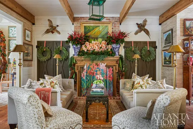 Living room with two neutral sofas and two patterned chaise lounges flanking a fireplace decorated for the holidays with wreaths, garlands, and floral arrangements.